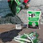 Close-up of a contractor pouring HP Asphalt Cold Patch into a pavement crack. Below, several bags of the cold patch are stacked near a completed pothole repair.'