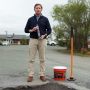 Man holding Tru Tack Cold Joint Sealer, standing with an asphalt crack in front of him and a concrete and asphalt patch with a tamper tool beside him.'